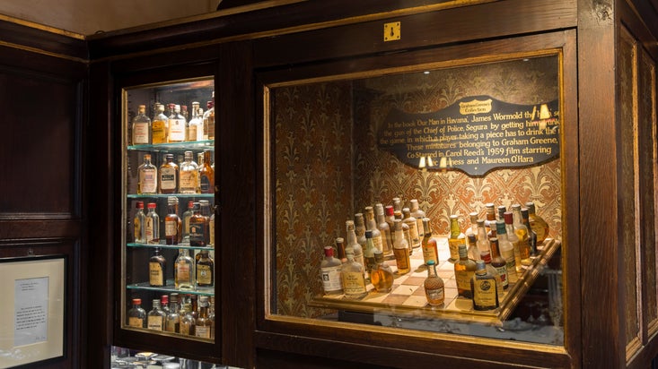 Museum-style display featuring a whisky-bottle chess set inspired by Our Man in Havana, with dozens of miniature spirits bottles arranged on a chessboard and illuminated shelves of vintage miniatures set against patterned wallpaper.